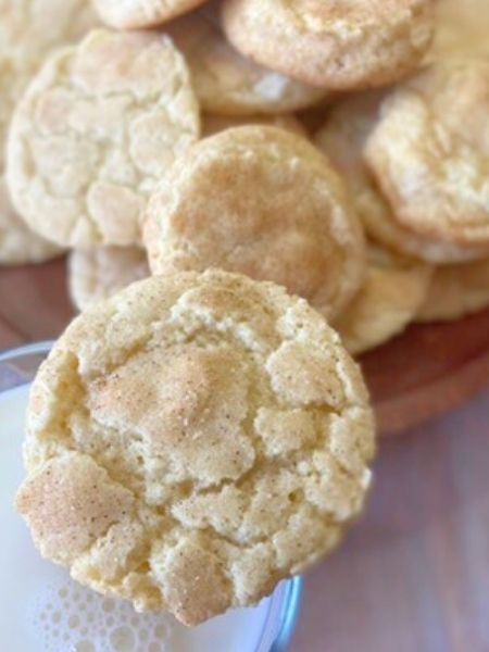 snickerdoodle cookies that are lightly golden and coated in cinnamon sugar sitting on a plate, one rests on a glass of milk